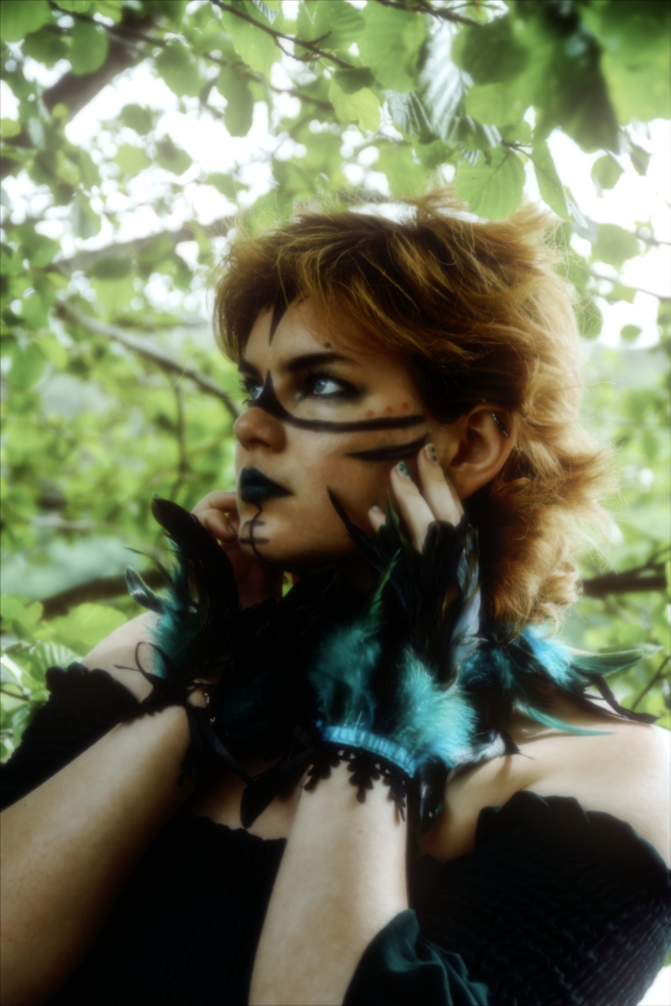 Portret kobiety z artystycznym makijażem w stylu leśnego ducha, z piórami i koronkowymi ozdobami na szyi, na tle rozmytej zieleni drzew.