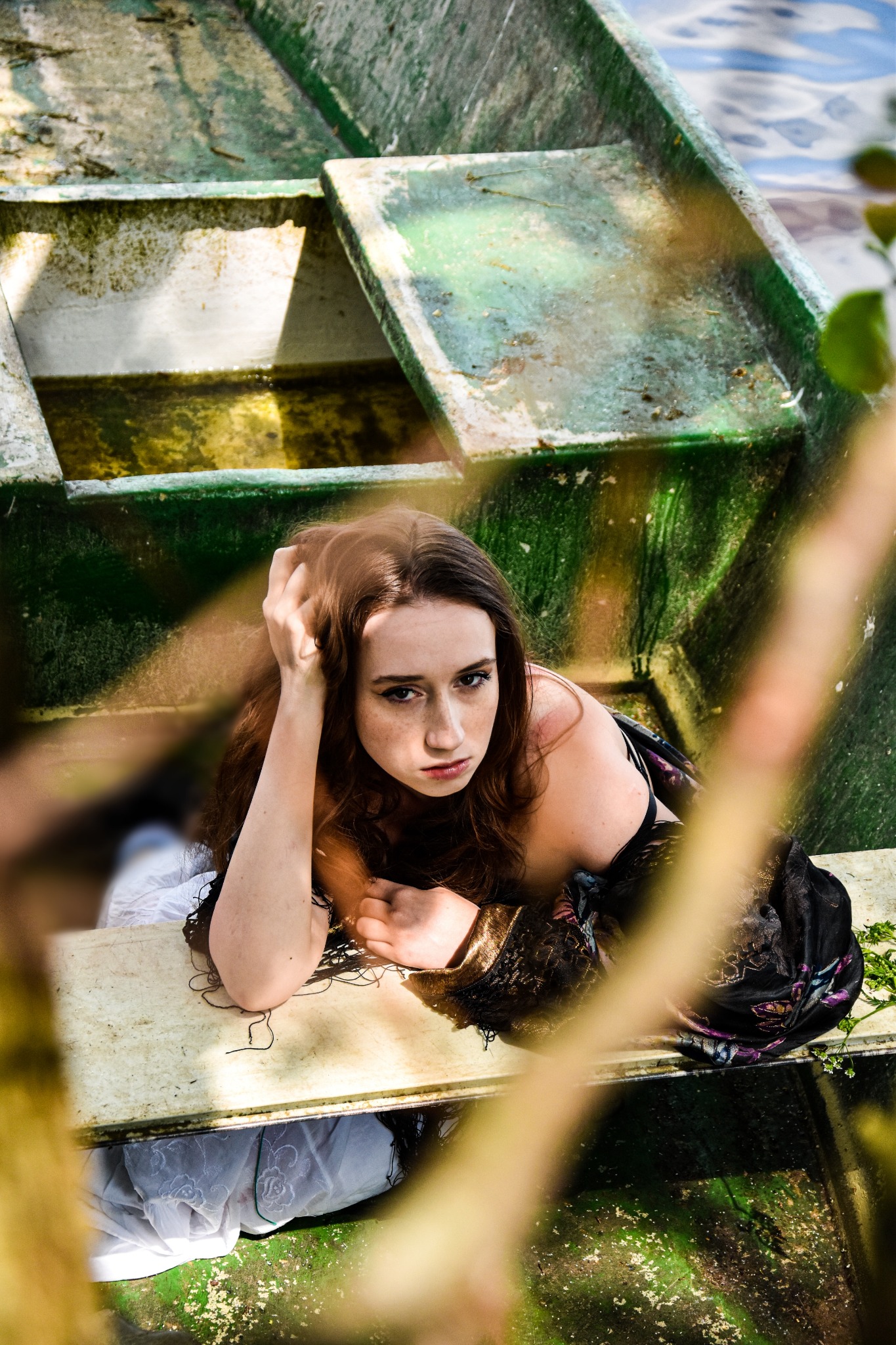 Portret kobiety z rudymi włosami leżącej w starej, zielonej łodzi z wodą, ujęcie zza gałęzi drzewa.
