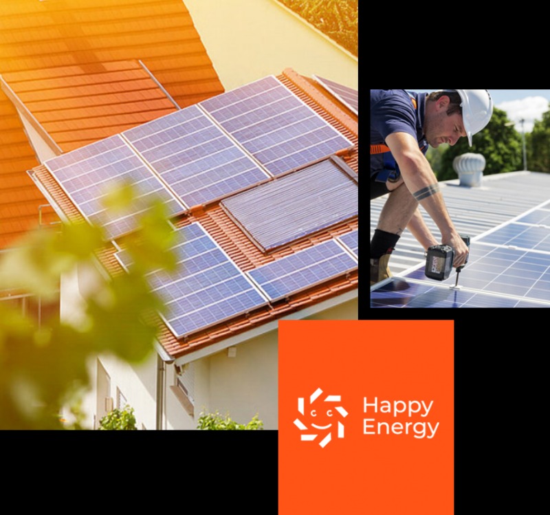 Montaż paneli słonecznych na dachu z czerwonej dachówki, widok z dołu na część budynku, instalator w kasku przykręca panel, pomarańczowe logo Happy Energy w prawym dolnym rogu.