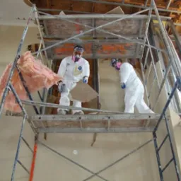 Dwóch pracowników w kombinezonach ochronnych na rusztowaniu podczas usuwania uszkodzonej ściany i różowej wełny mineralnej w pomieszczeniu w trakcie osuszania.