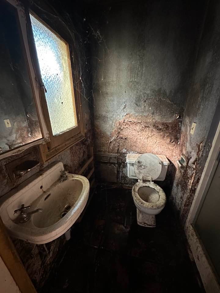 Zniszczona łazienka po zalaniu lub pożarze: brudne ściany z zaciekami, pajęczyny w rogu przy oknie, stara umywalka i muszla klozetowa pokryte osadem.