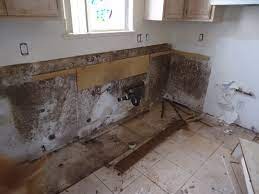 Zniszczona ściana i podłoga w kuchni po zalaniu, widoczne ślady wilgoci i pleśni, brakujące elementy wyposażenia, odsłonięte rury, fragment deski na podłodze.