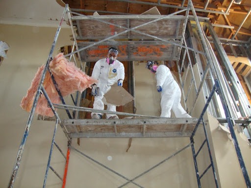 Dwóch pracowników w kombinezonach ochronnych na rusztowaniu podczas usuwania uszkodzonej ściany i różowej wełny mineralnej w pomieszczeniu w trakcie osuszania.