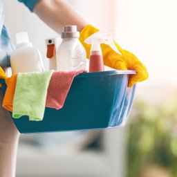 Blysk - Osoba w żółtych rękawicach trzyma niebieski pojemnik z butelkami środków czystości, ściereczkami z mikrofibry w kolorach pomarańczowym i zielonym, w tle rozproszone światło i zieleń roślin.