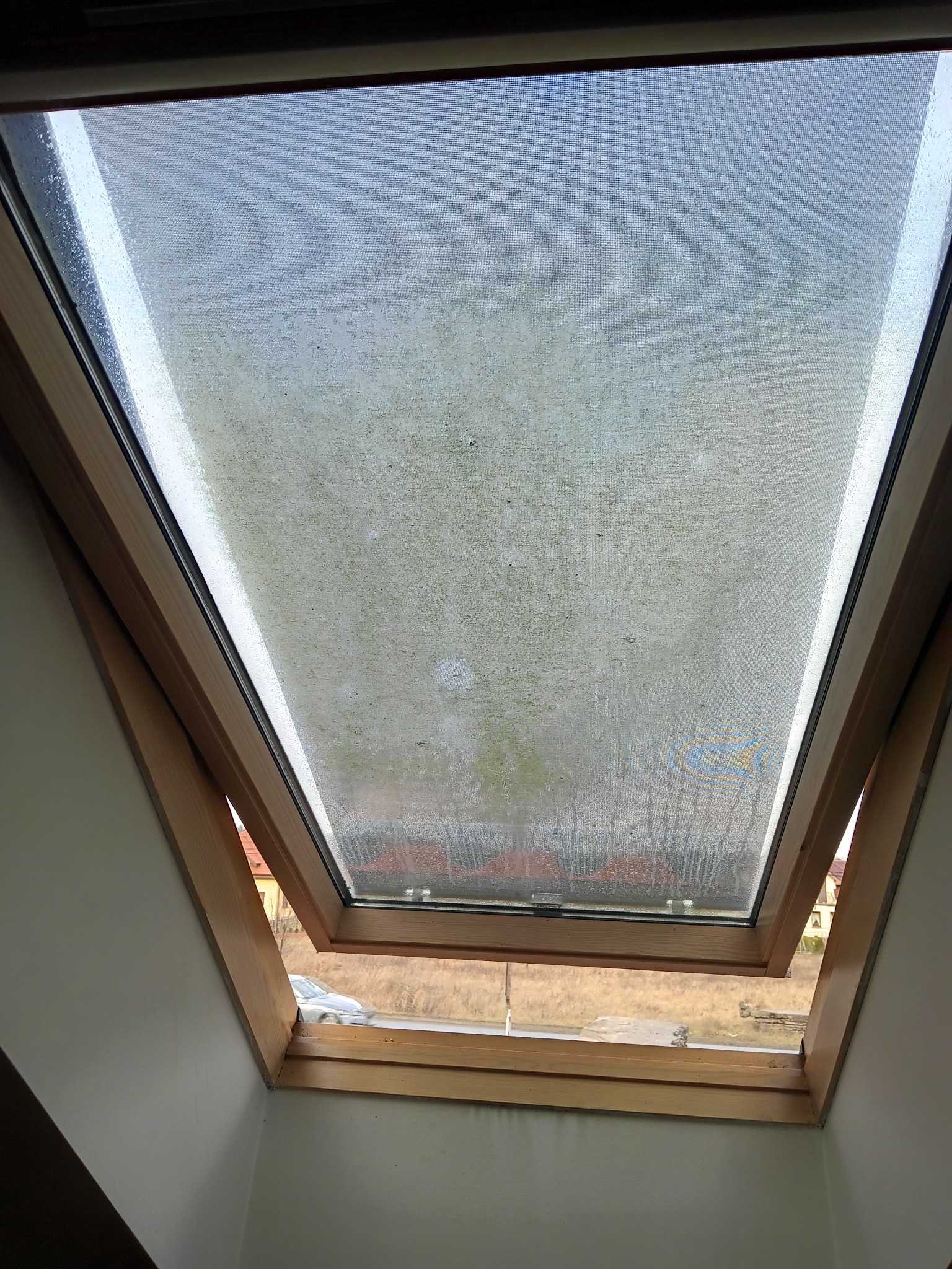 Brudne okno dachowe z drewnianą ramą, widoczne zanieczyszczenia i zacieki na szybie, przez którą widać fragment ulicy i przejeżdżający samochód.