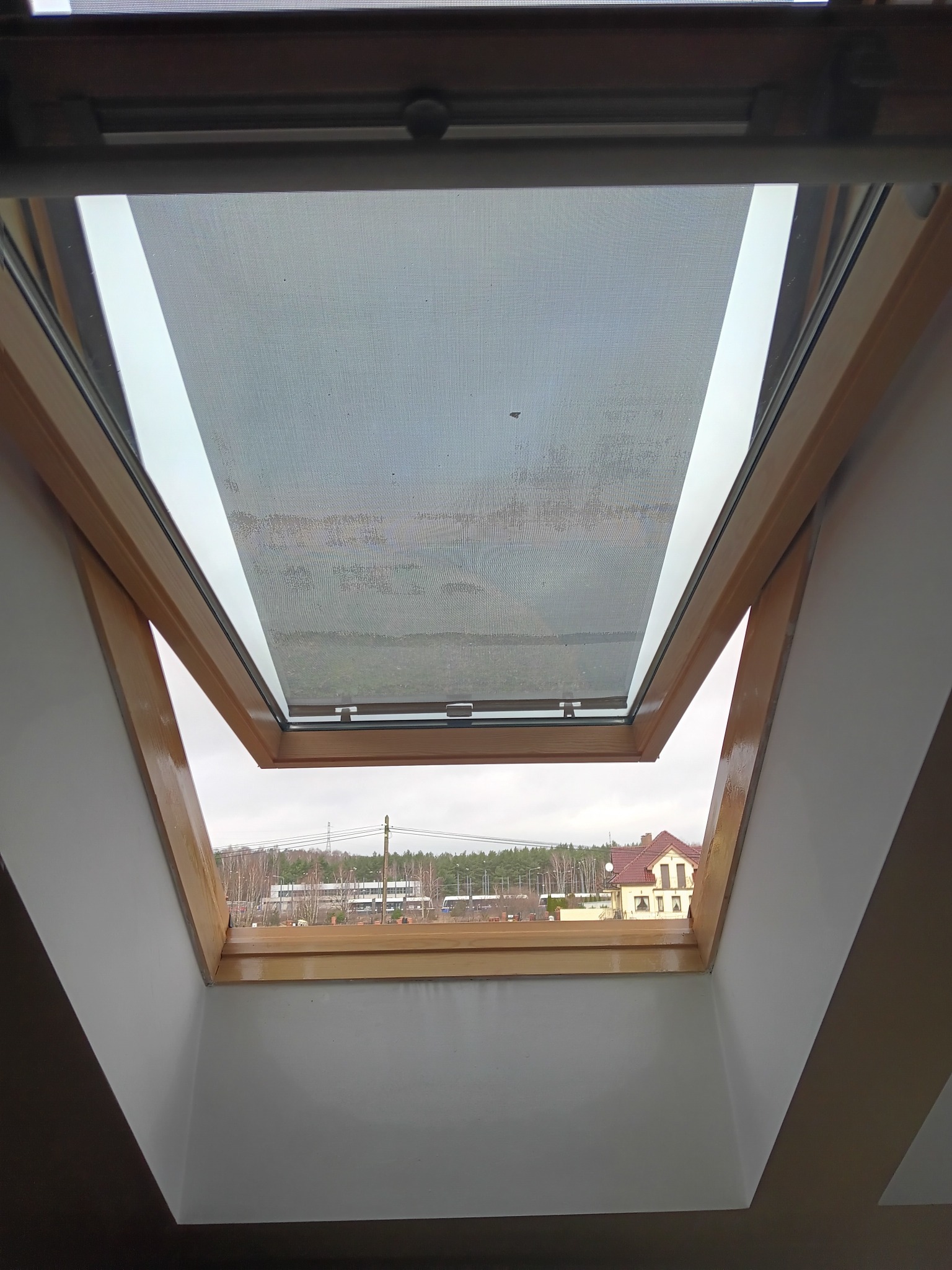 Otwarty drewniany okno dachowe z widokiem na budynki i drzewa w pochmurny dzień.