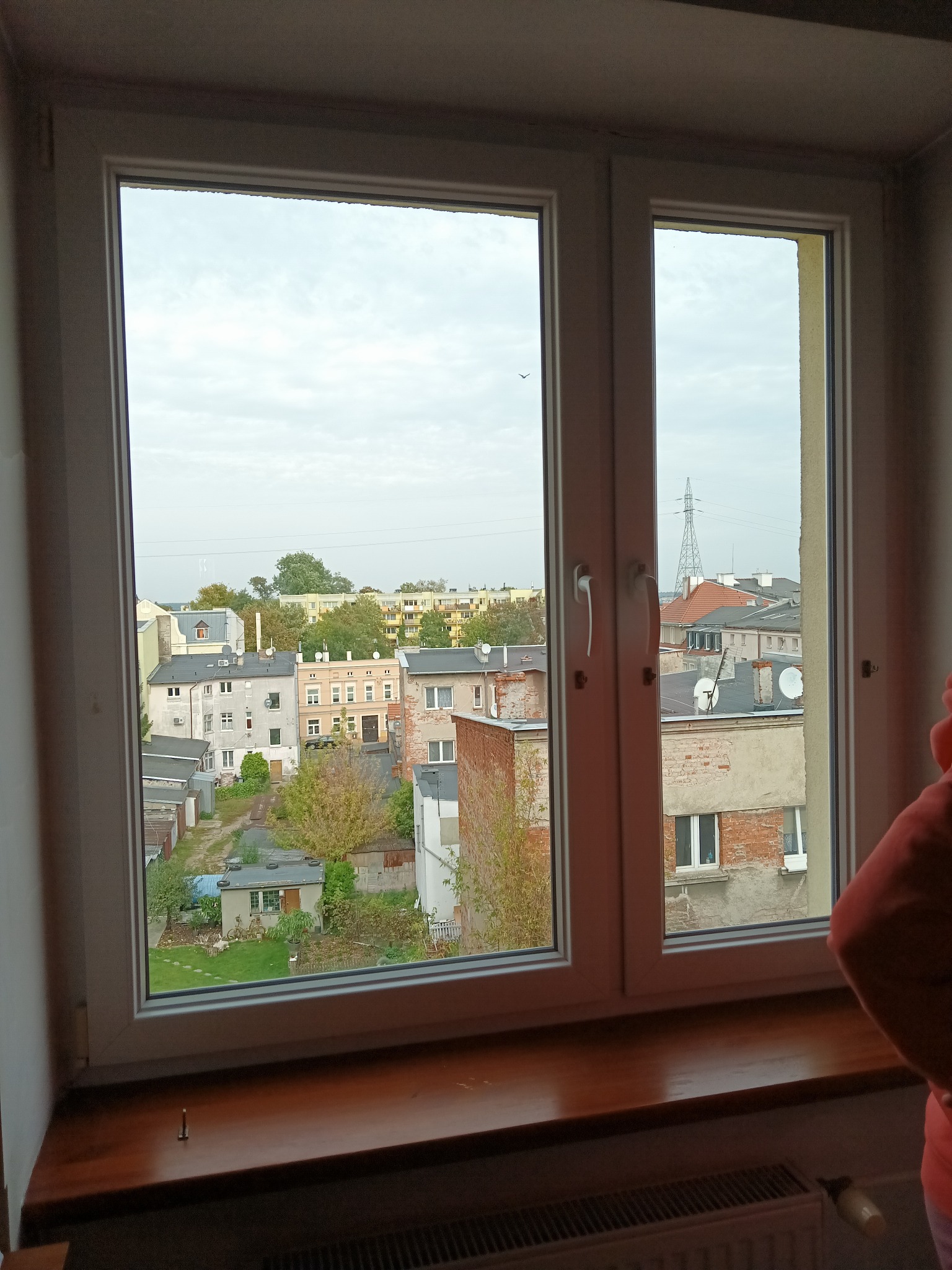 Widok z okna PCV na zabudowę miejską z zielenią, widoczna sieć energetyczna w tle, okno czyste, ale z lekkimi śladami po kroplach deszczu.