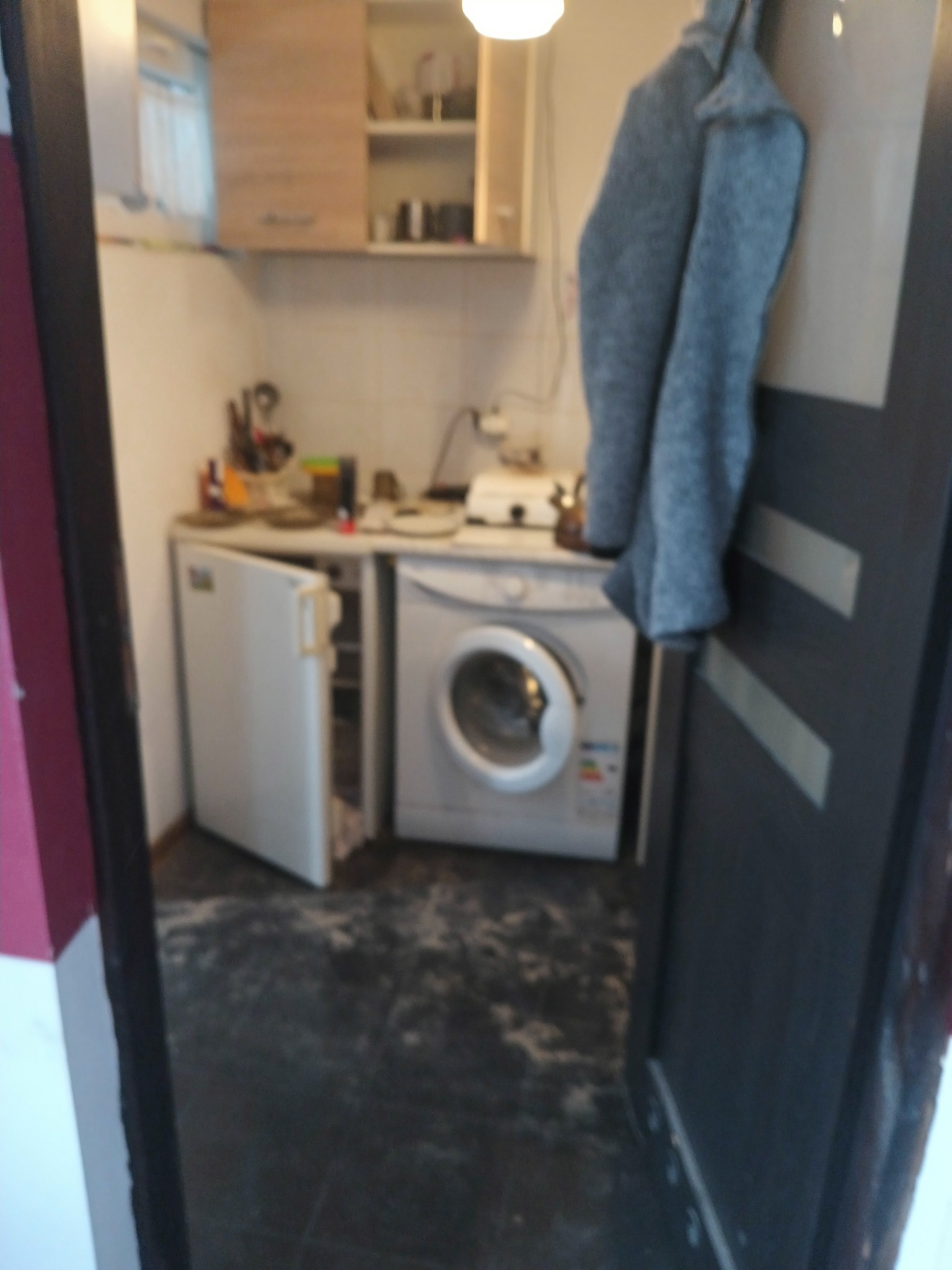 Nieduża kuchnia z otwartą lodówką i pralką, widok zza uchylonych drzwi, szara kurtka wisi na drzwiach.