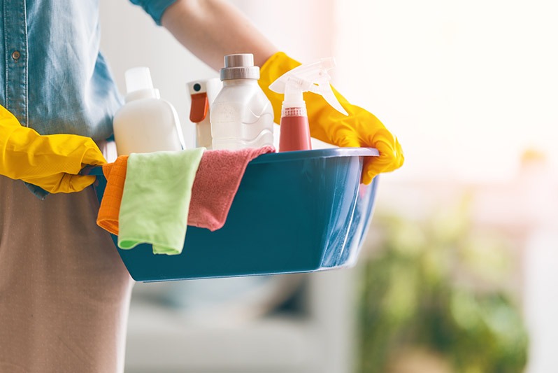 Osoba w żółtych rękawicach trzyma niebieski pojemnik z butelkami środków czystości, ściereczkami z mikrofibry w kolorach pomarańczowym i zielonym, w tle rozproszone światło i zieleń roślin.