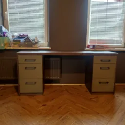 Biurko z szufladami w odcieniach brązu i zieleni, ustawione pod oknami z żaluzjami, na parkiecie ułożonym w jodełkę.