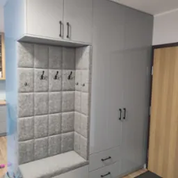 Szara szafa wnękowa z siedziskiem i tapicerowanym panelem ściennym z haczykami, czarne minimalistyczne uchwyty, widok z przedsionka.