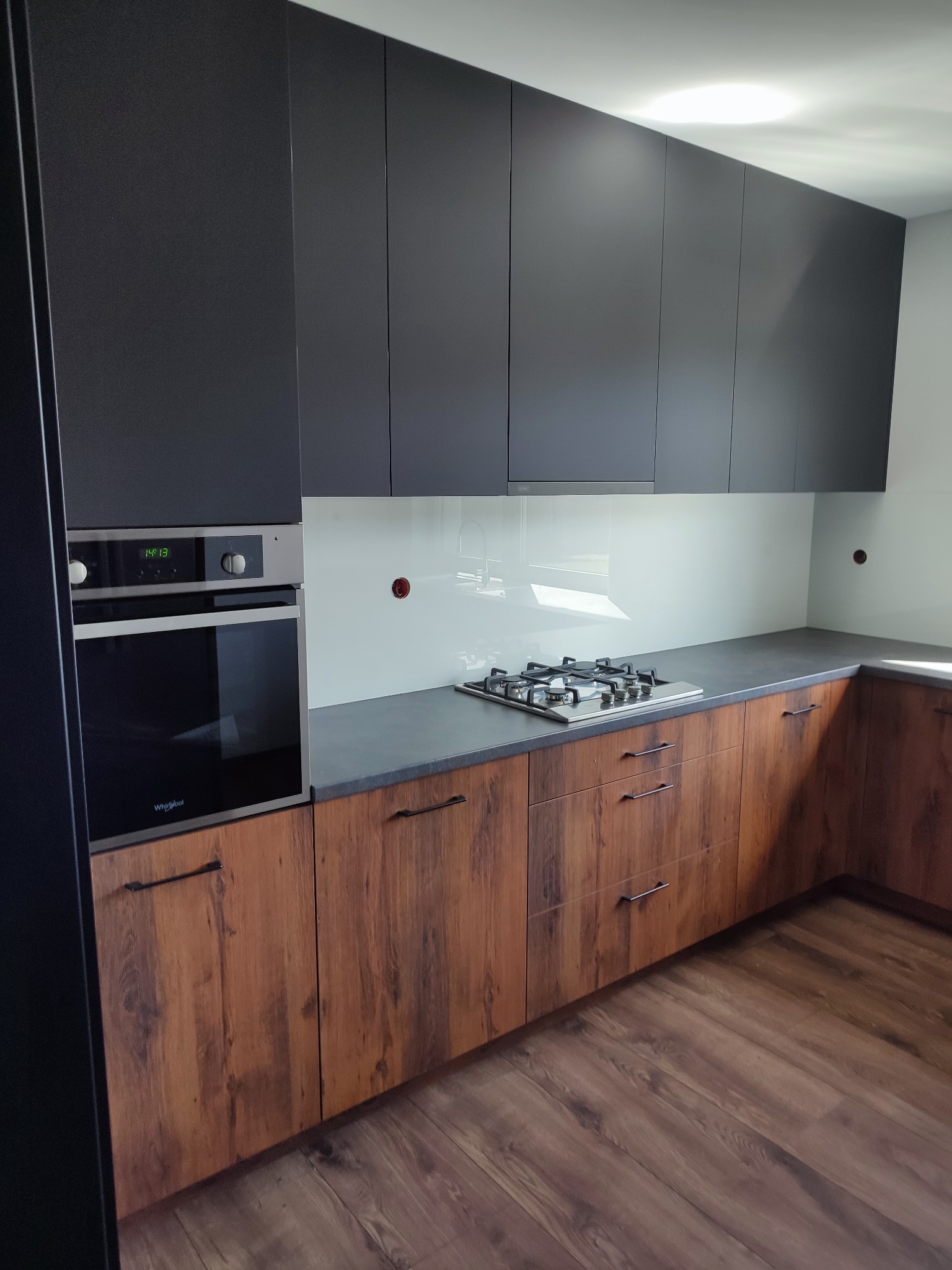 Nowoczesna kuchnia z ciemnymi górnymi szafkami, drewnopodobnymi dolnymi szafkami i szklanym panelem nad blatem, widoczna płyta gazowa i piekarnik Whirlpool.