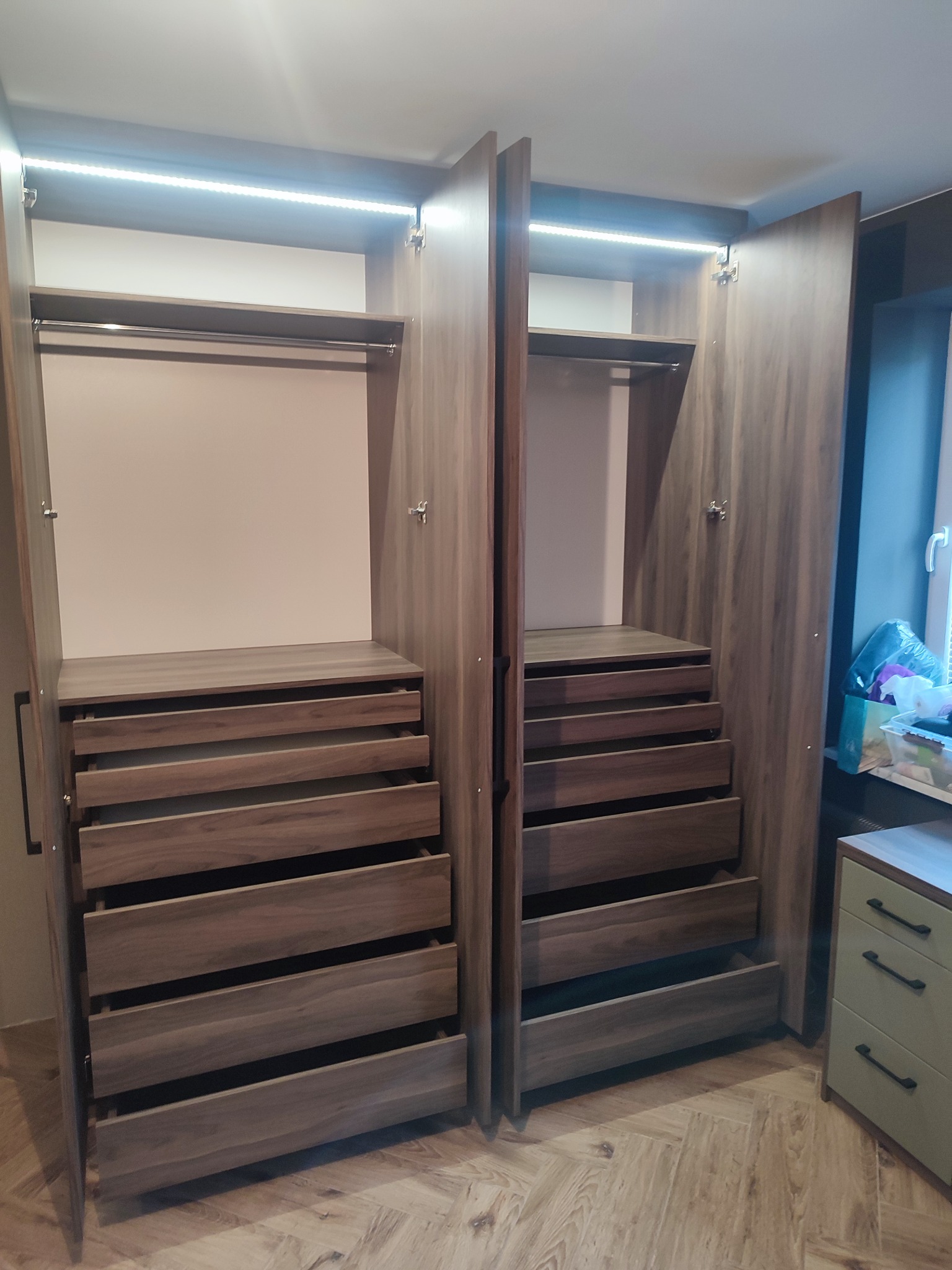 Wnętrze szafy wnękowej z otwartymi drzwiami, widoczne półki i szuflady z drewna o naturalnym odcieniu, oświetlenie LED zamontowane u góry, na podłodze parkiet w jodełkę.