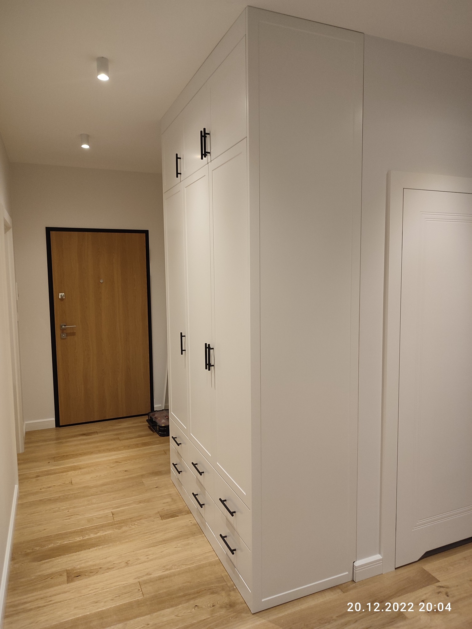 Biała szafa wnękowa z czarnymi uchwytami w przedpokoju z drewnianą podłogą, widoczna z perspektywy podłogi, obok drzwi wejściowych.