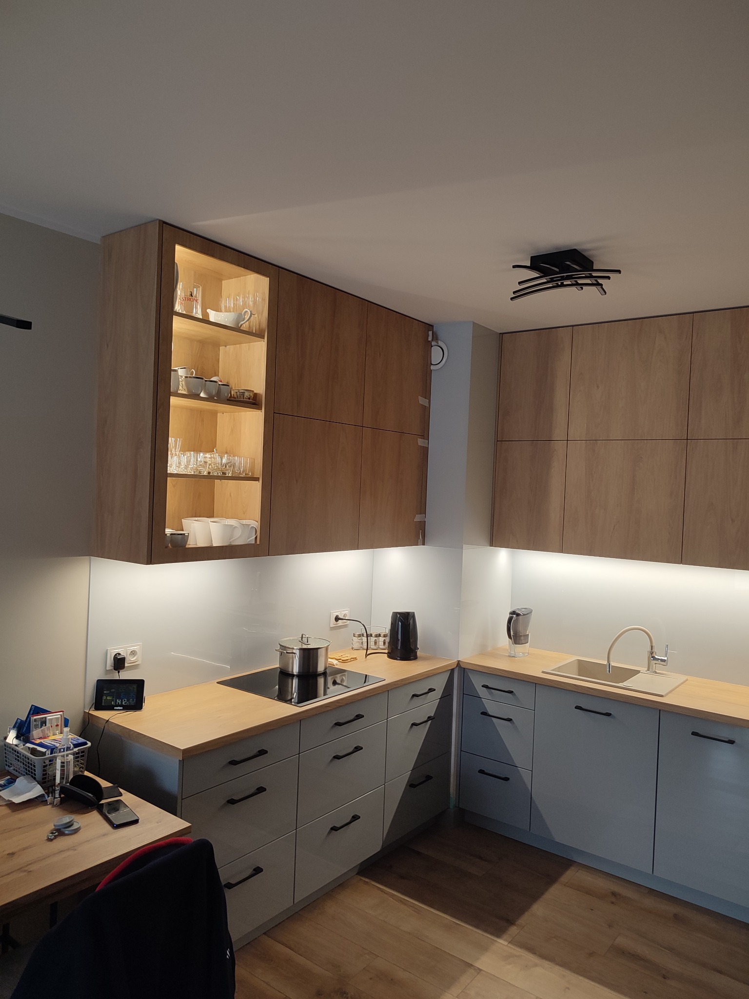 Nowoczesna kuchnia z drewnianymi szafkami górnymi z podświetleniem i szarymi szafkami dolnymi, drewniany blat, czarna płyta indukcyjna z garnkiem, minimalistyczna lampa sufitowa.