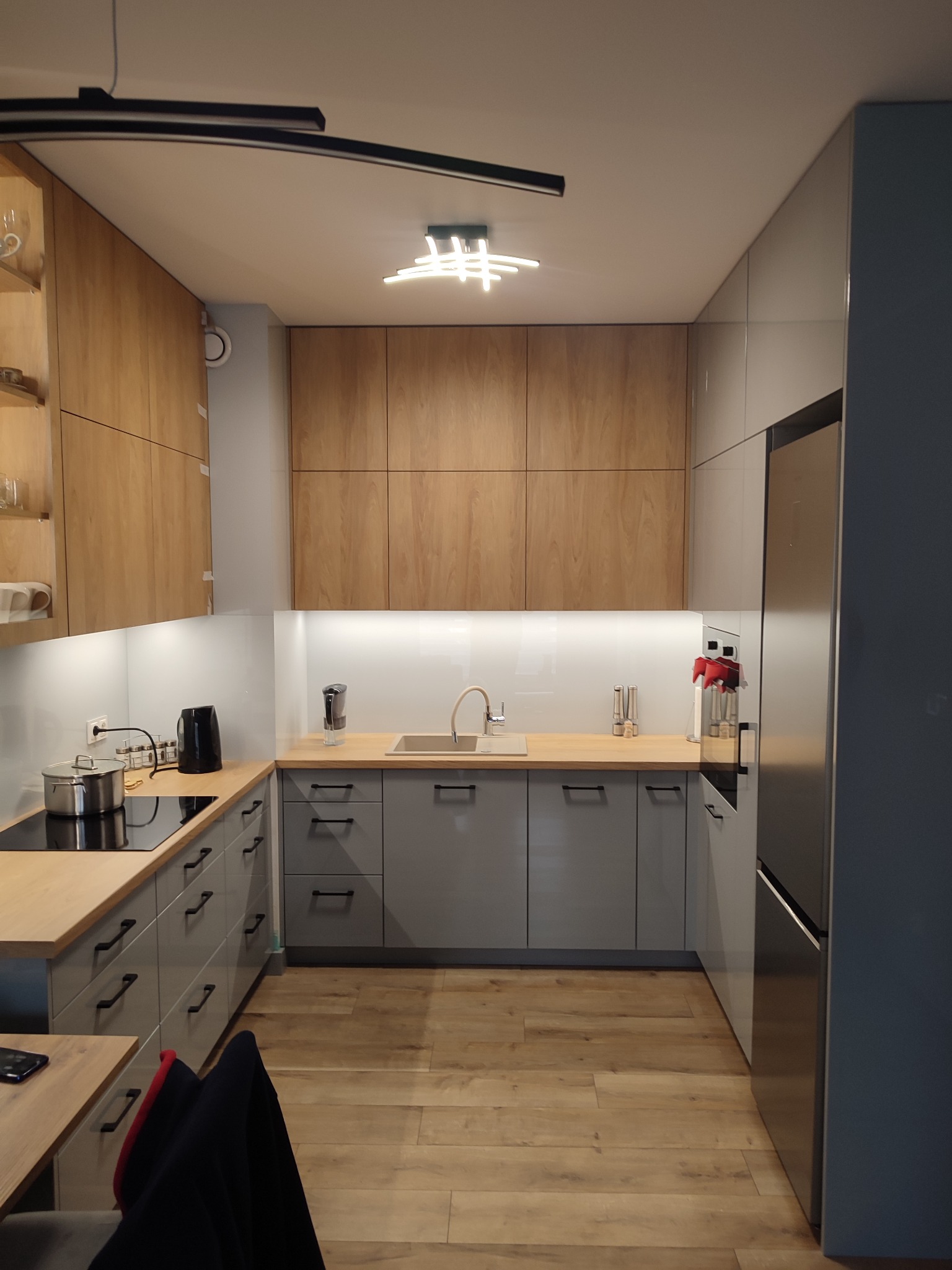 Nowoczesna kuchnia w odcieniach szarości i drewna, z drewnianą podłogą i oryginalnym oświetleniem sufitowym, widok na blaty, szafki i sprzęt AGD.