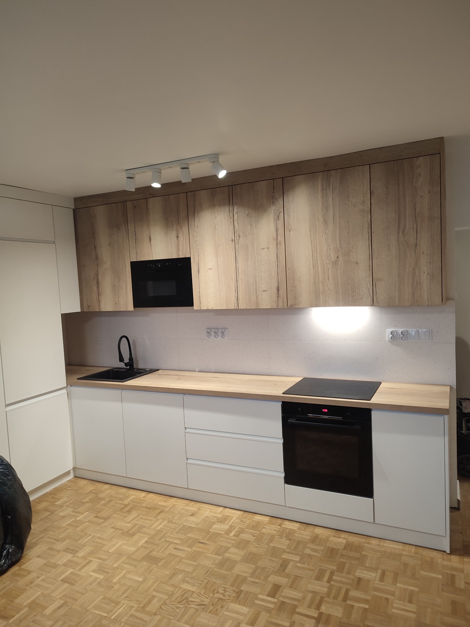 Nowoczesna kuchnia z białymi dolnymi szafkami i górnymi szafkami z forniru drewnopodobnego, czarnym zlewem i piekarnikiem, oświetlona reflektorami sufitowymi, na parkiecie w jodełkę.