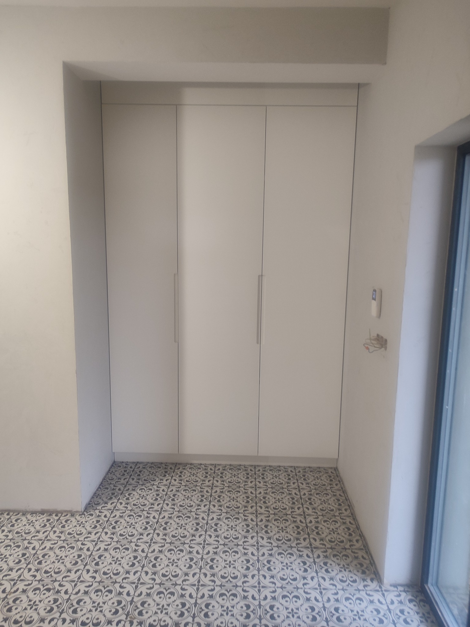 Trzydrzwiowa szafa wnękowa z białymi frontami i minimalistycznymi uchwytami, wbudowana w ścianę, na podłodze wzorzyste płytki.