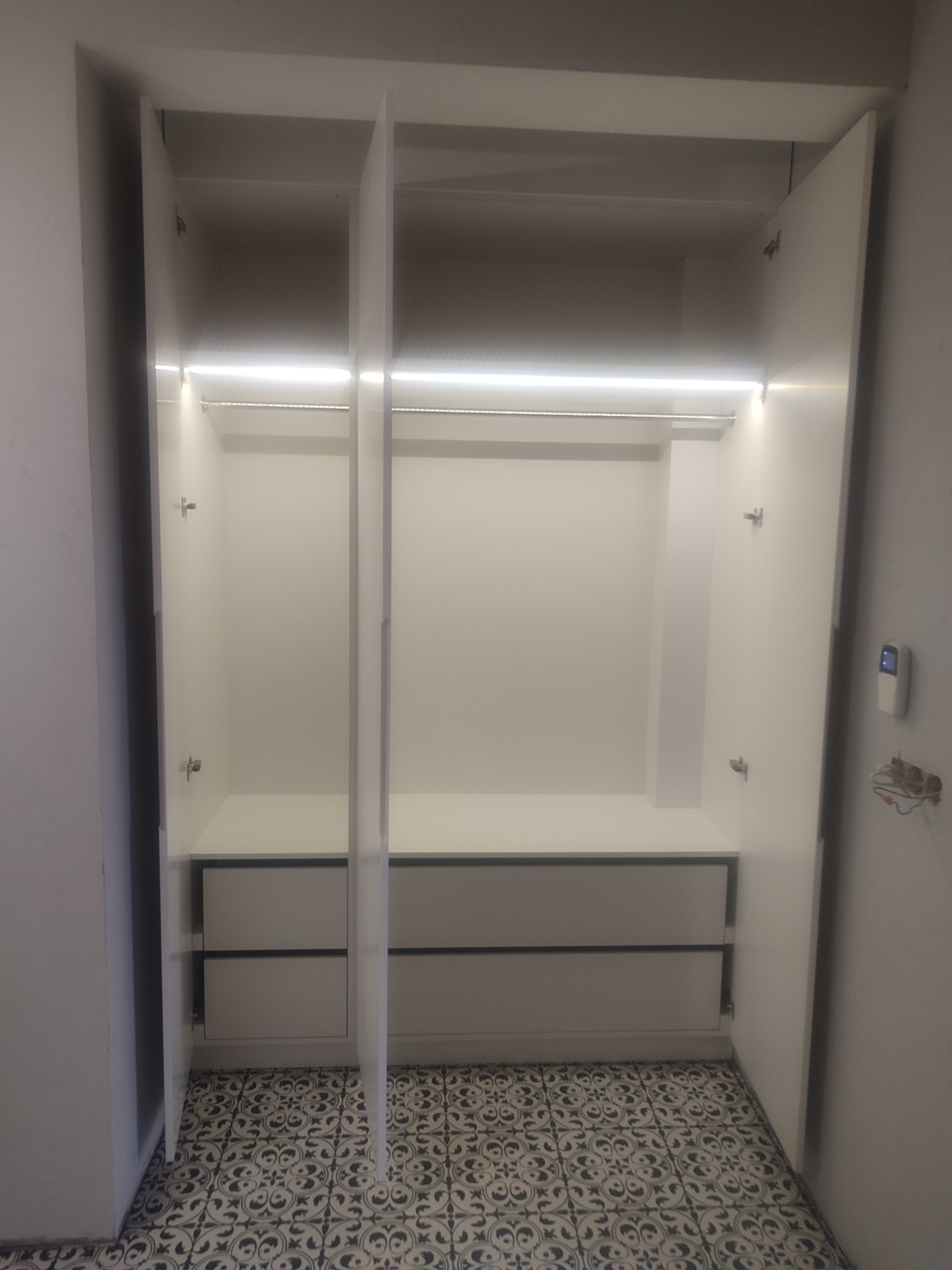 Wnętrze szafy wnękowej z oświetleniem LED, widoczne półki, szuflady i drążek na ubrania, na podłodze wzorzysta płytka.