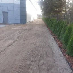 Przygotowany teren pod trawnik z widocznymi śladami po maszynach, obok rząd świeżo posadzonych tuj i metalowe ogrodzenie.