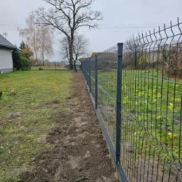 Nowo postawione, panelowe ogrodzenie w kolorze grafitowym, oddzielające trawnik od świeżo przekopanej ziemi, z drzewami w tle i fragmentem domu po lewej stronie, uchwycone w pochmurny dzień.