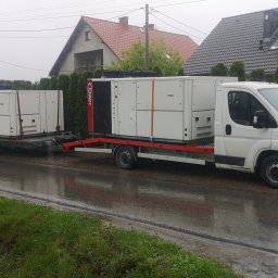 V.I.P-TRANS - Biały bus z przyczepą transportuje dwie duże, białe jednostki klimatyzacyjne marki Lennox na mokrej, asfaltowej drodze w deszczowy dzień. W tle widoczne domy jednorodzinne z ogrodami.