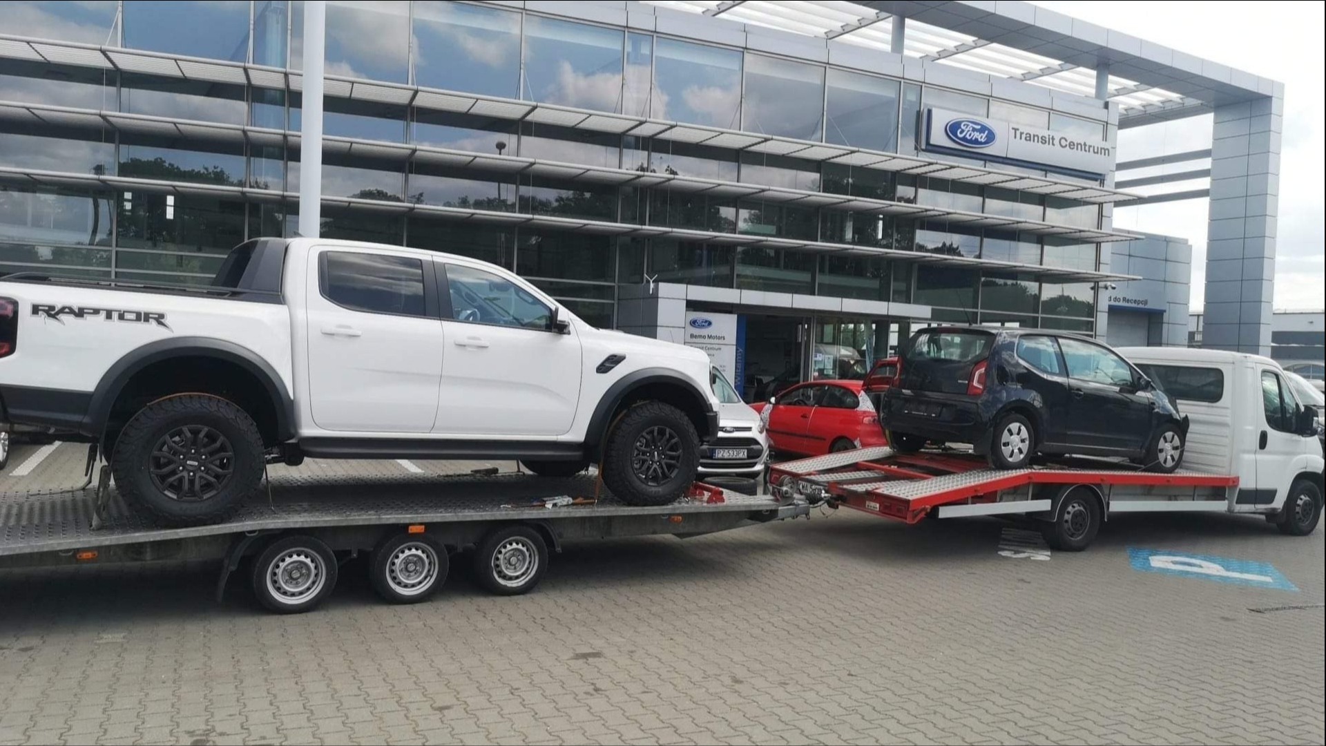 Biały Ford Ranger Raptor na niskopodwoziowej lawecie obok transportu busa z czarnym samochodem osobowym na tle salonu Ford Transit Centrum.