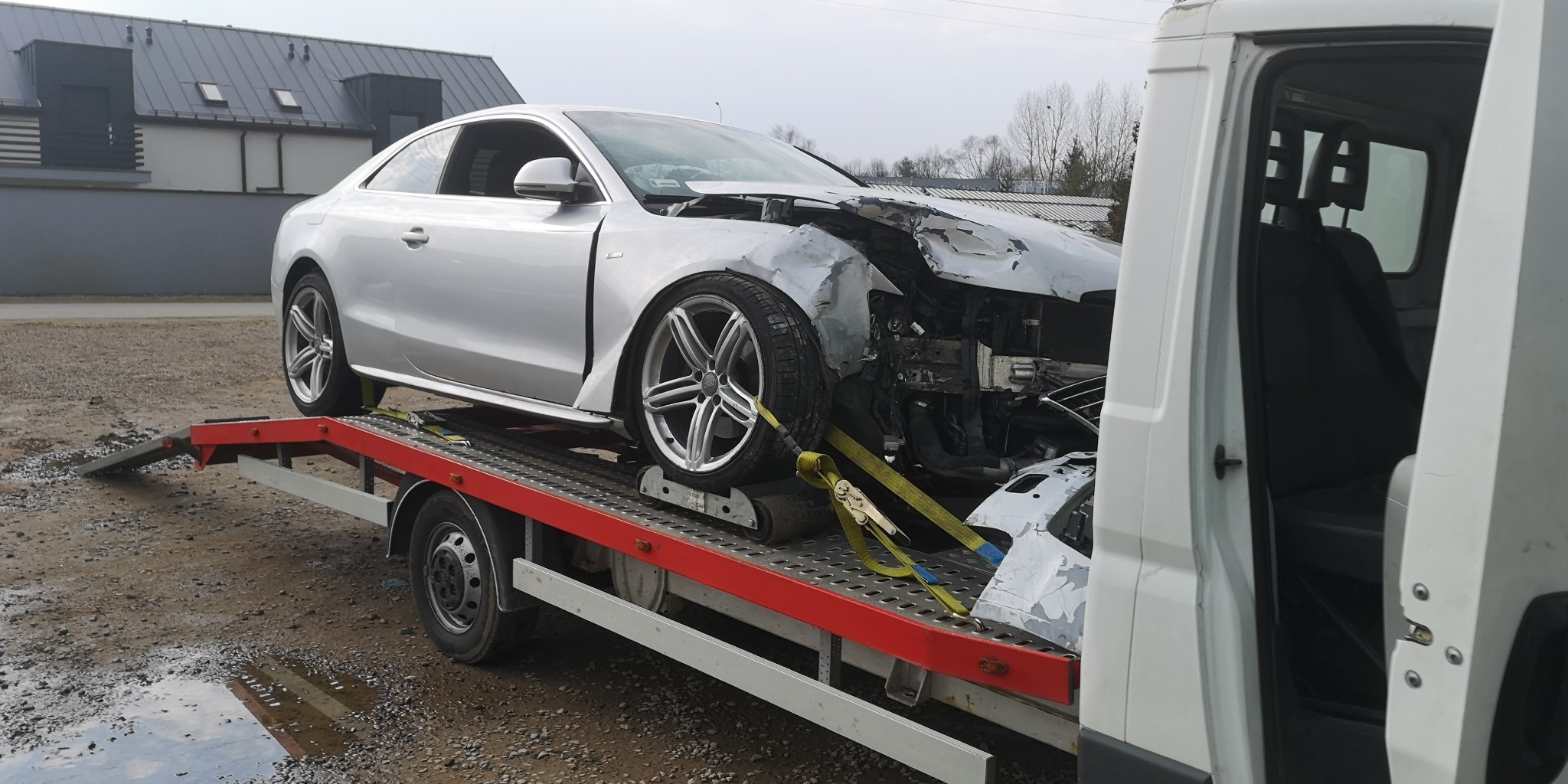 Srebrne Audi A5 z uszkodzonym przodem, zabezpieczone pasami, na czerwonej lawecie z najazdami, widok z boku na tle szarego budynku i białego busa.