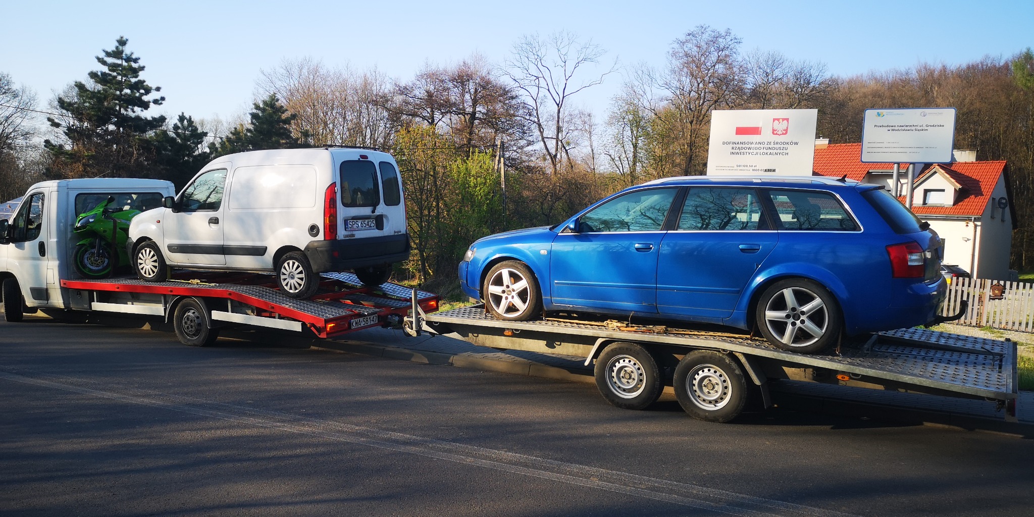 Dwa samochody i motocykl transportowane na lawetach; biały furgon i zielony motocykl na jednej, niebieskie kombi na drugiej; w tle tablice informacyjne i drzewa.