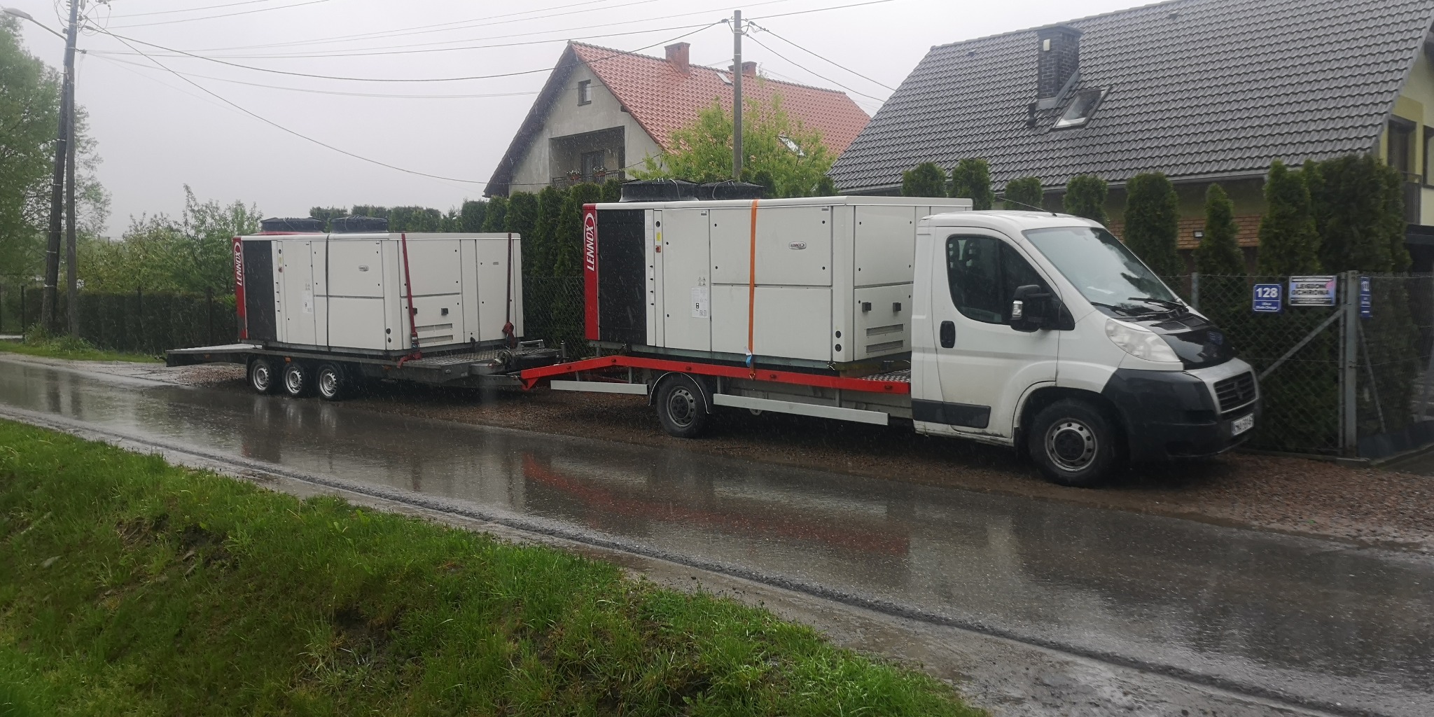 Biały bus z przyczepą transportuje dwie duże, białe jednostki klimatyzacyjne marki Lennox na mokrej, asfaltowej drodze w deszczowy dzień. W tle widoczne domy jednorodzinne z ogrodami.