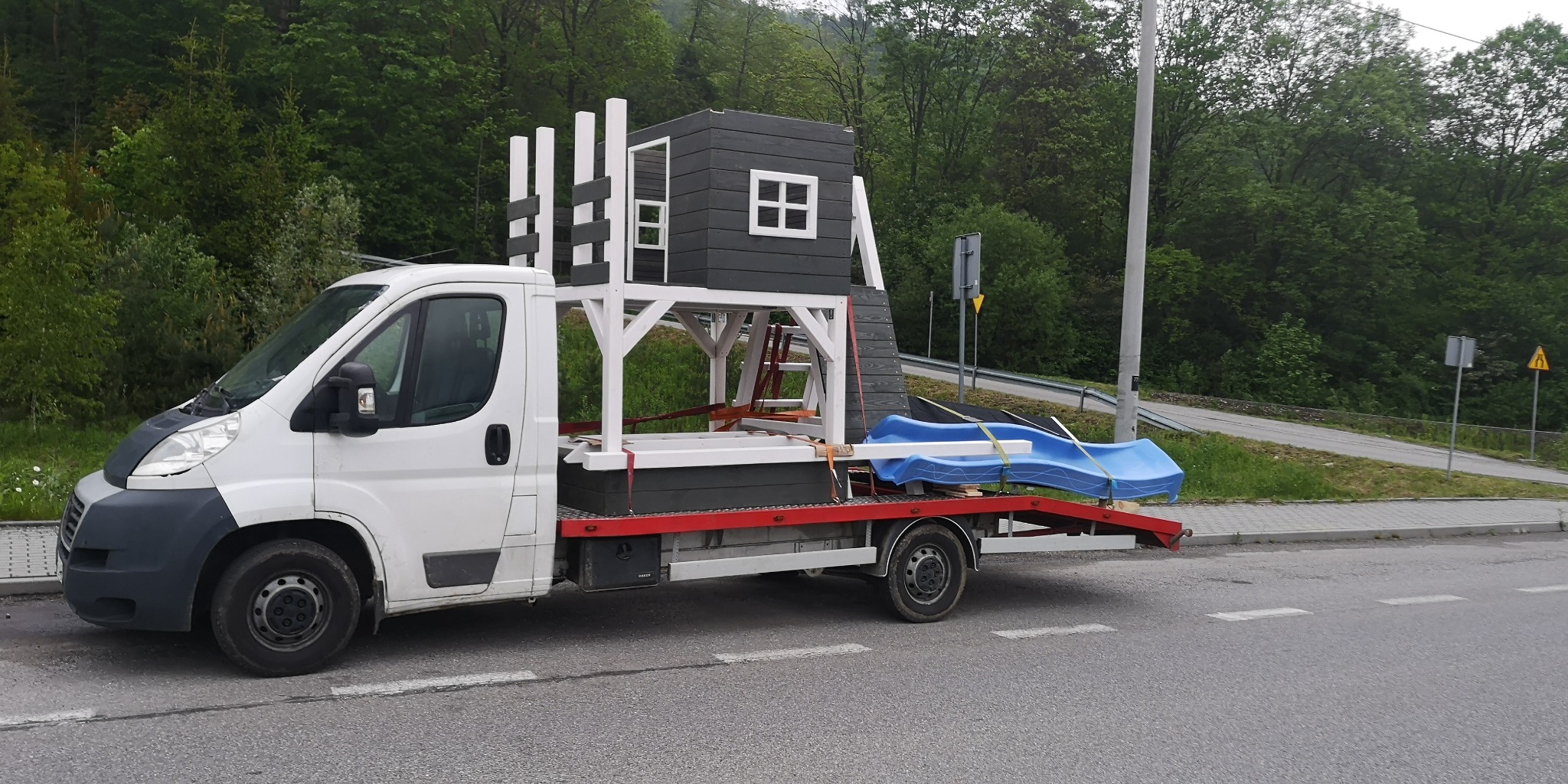 Biała laweta przewozi szarą drewnianą konstrukcję domku zabaw z białą ramą i niebieską zjeżdżalnią na tle zieleni.