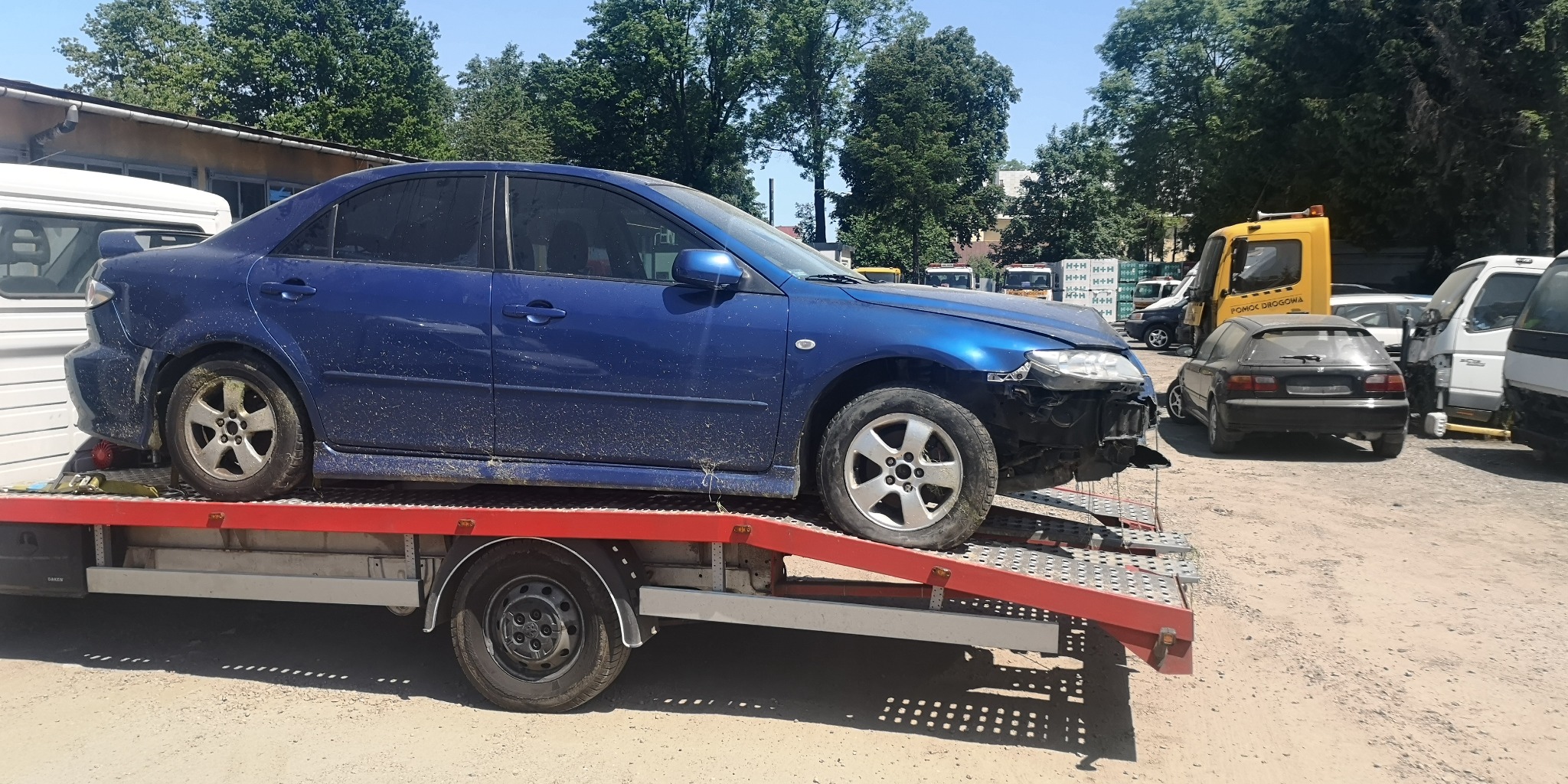 Uszkodzony, niebieski samochód osobowy marki Mazda, przewożony na czerwonej platformie lawety w słoneczny dzień, z widocznym żółtym pojazdem pomocy drogowej w tle.