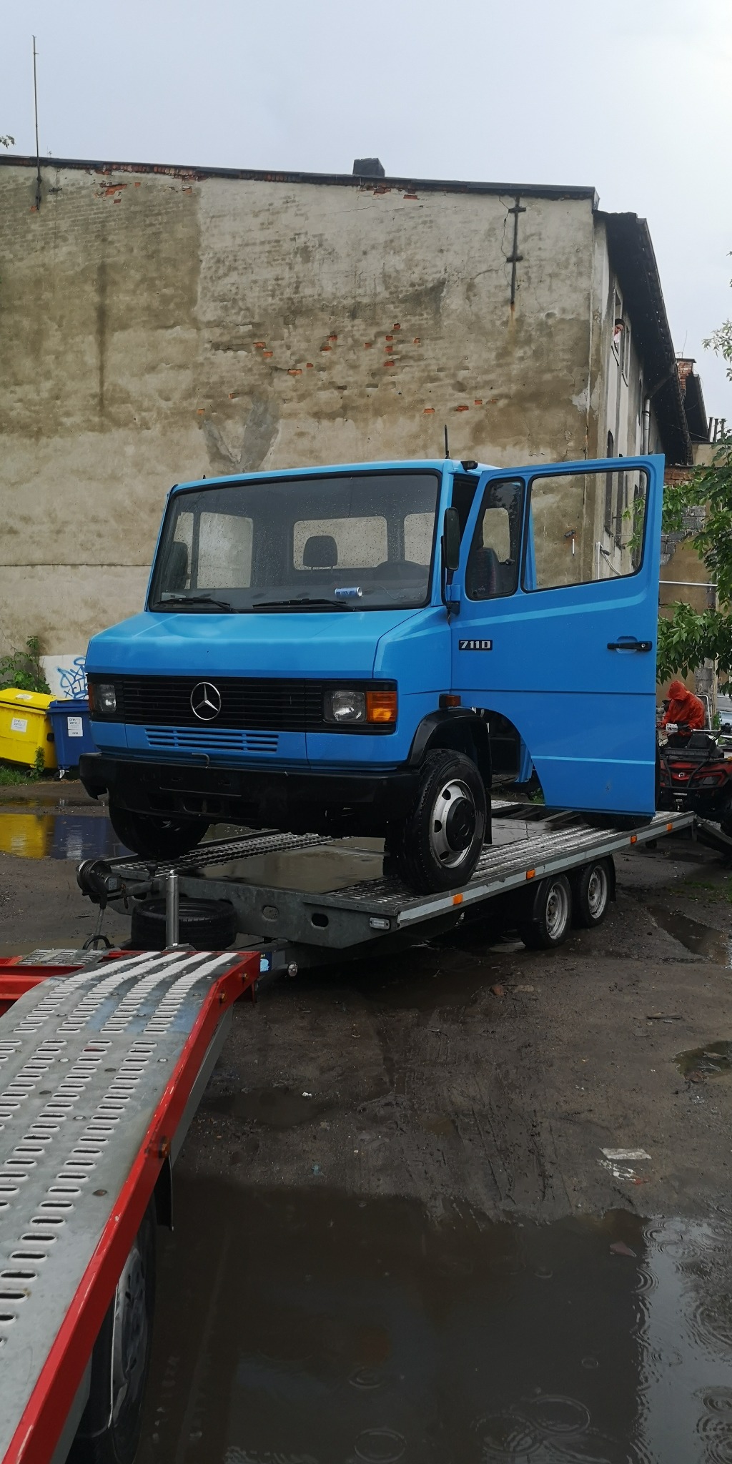 Niebieski Mercedes 711D na platformie lawety z otwartymi drzwiami, na tle starego budynku w deszczowy dzień.