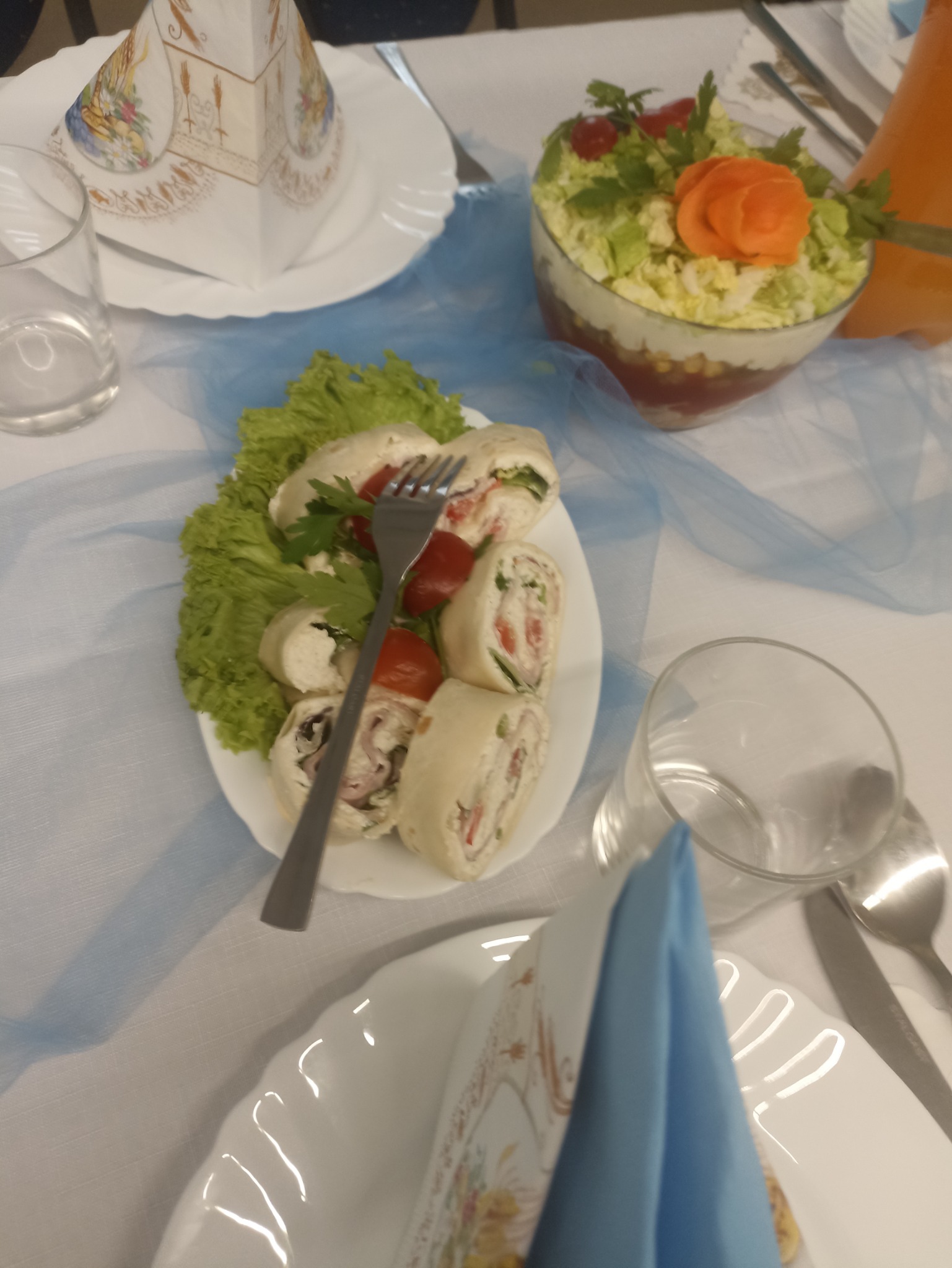 Dekoracja stołu z roladkami z szynką, sałatą i pomidorami, sałatką warzywną z marchewkową rozetą i szklankami na białym obrusie z niebieską dekoracyjną wstążką.
