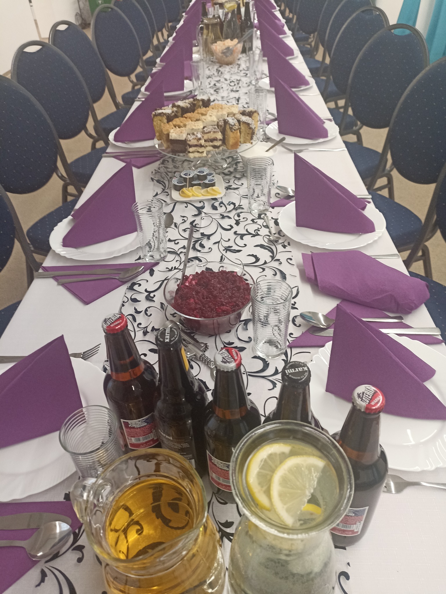 Ustawiony stół na imprezę: biały obrus w czarne wzory, fioletowe serwetki, szklanki, talerze, sztućce, ciasta na paterze, miska z sałatką, dzbanek z napojem i butelki z piwem.