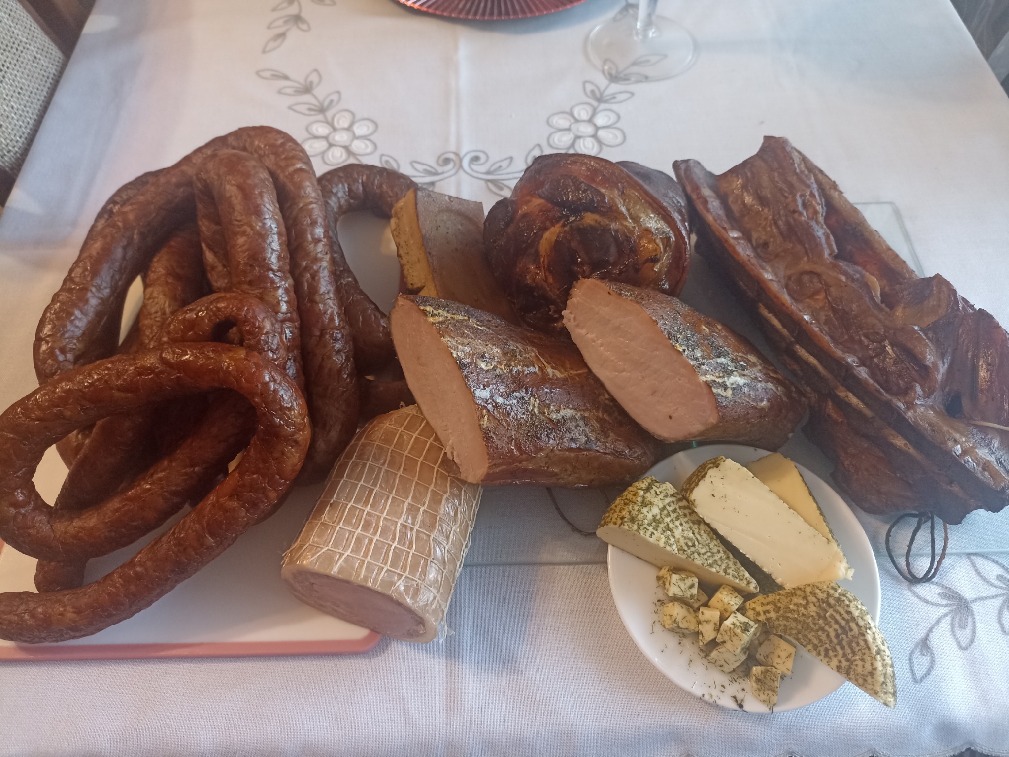 Kompozycja wędlin i serów na białym obrusie w kwiaty, widoczne kiełbasy, szynka w siatce, wędzonka i ser z ziołami na talerzyku.