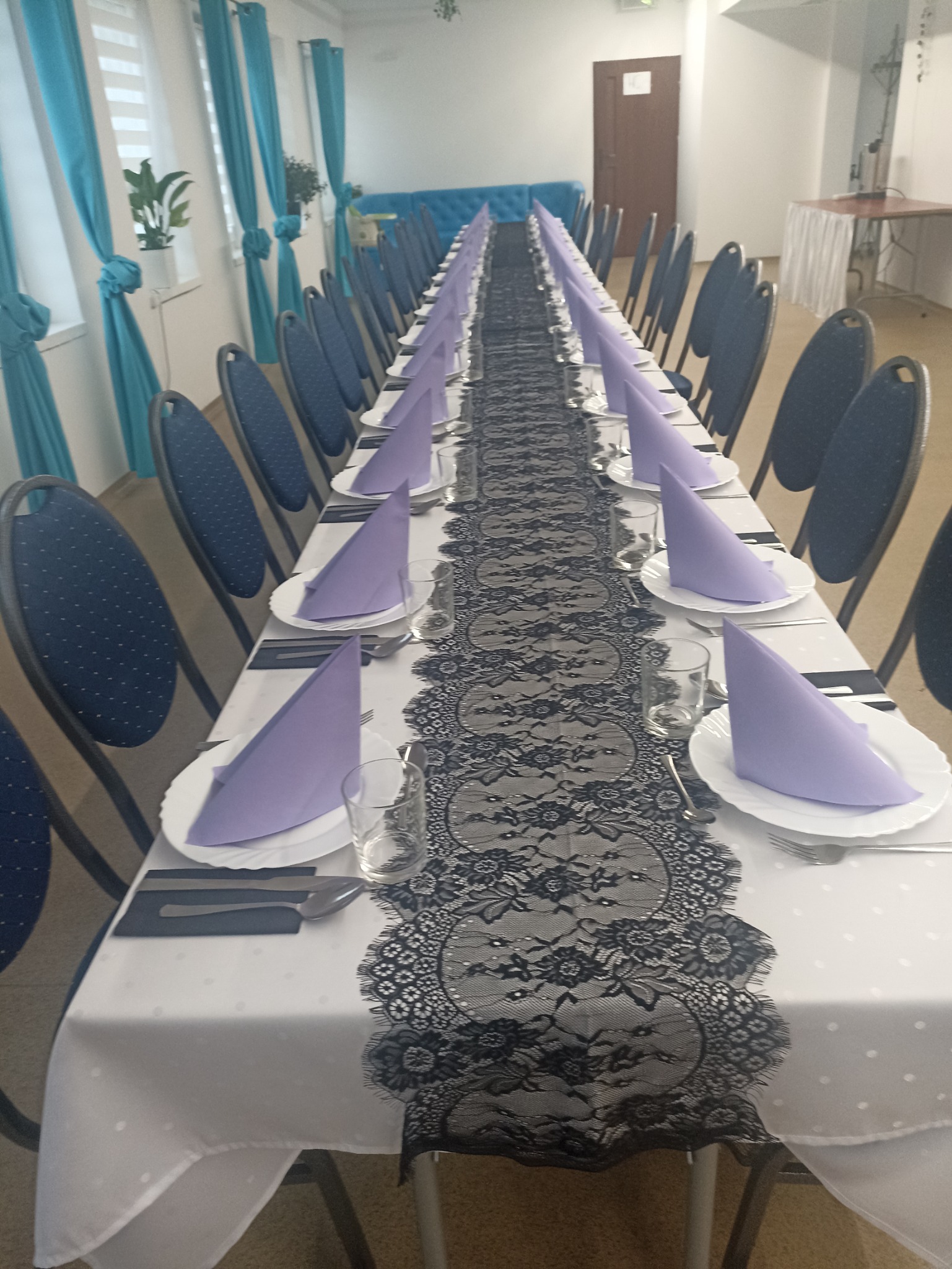 Długi stół nakryty białym obrusem w kropki, z czarnym koronkowym bieżnikiem, talerzami, sztućcami, szklankami i liliowymi serwetkami. Krzesła z granatową tapicerką ustawione wzdłuż stołu. W tle...