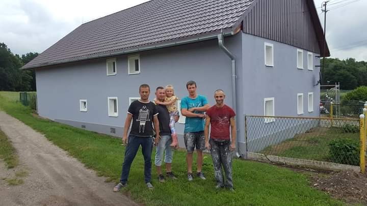 Grupa pięciu osób, w tym dziecko, pozuje na tle świeżo pomalowanego, dwupiętrowego domu z brązowym dachem i szarymi ścianami. Jeden z mężczyzn ma spodnie popryskane farbą.