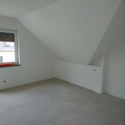 ZTM - Poddasze po remoncie, świeżo pomalowane na biało, widoczne gładkie ściany i sufit, szara wylewka na podłodze, okno z roletą, elementy instalacji elektrycznej na ścianie.