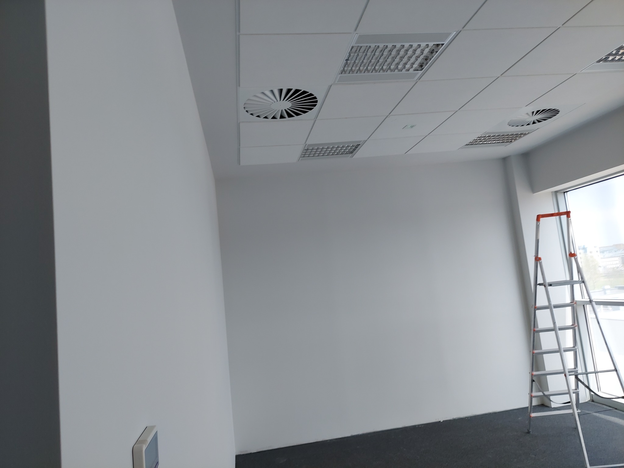 Świeżo pomalowane, jasnoszare ściany biura z widocznym sufitem kasetonowym i kratkami wentylacyjnymi, drabina oparta o ścianę przy oknie.