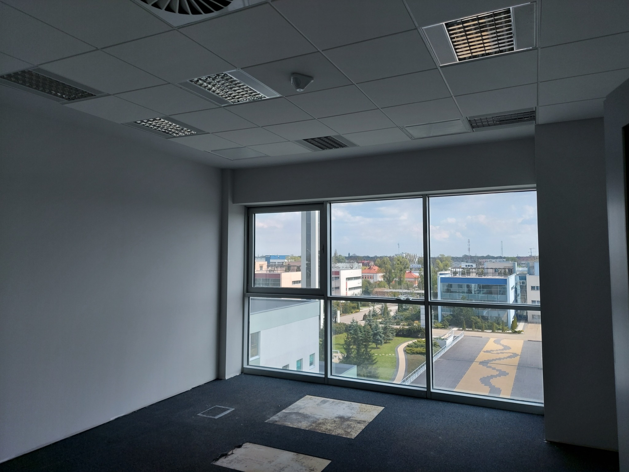 Puste biuro z wykładziną dywanową, widokiem na miasto przez duże okna i kasetonowym sufitem z oświetleniem rastrowym.