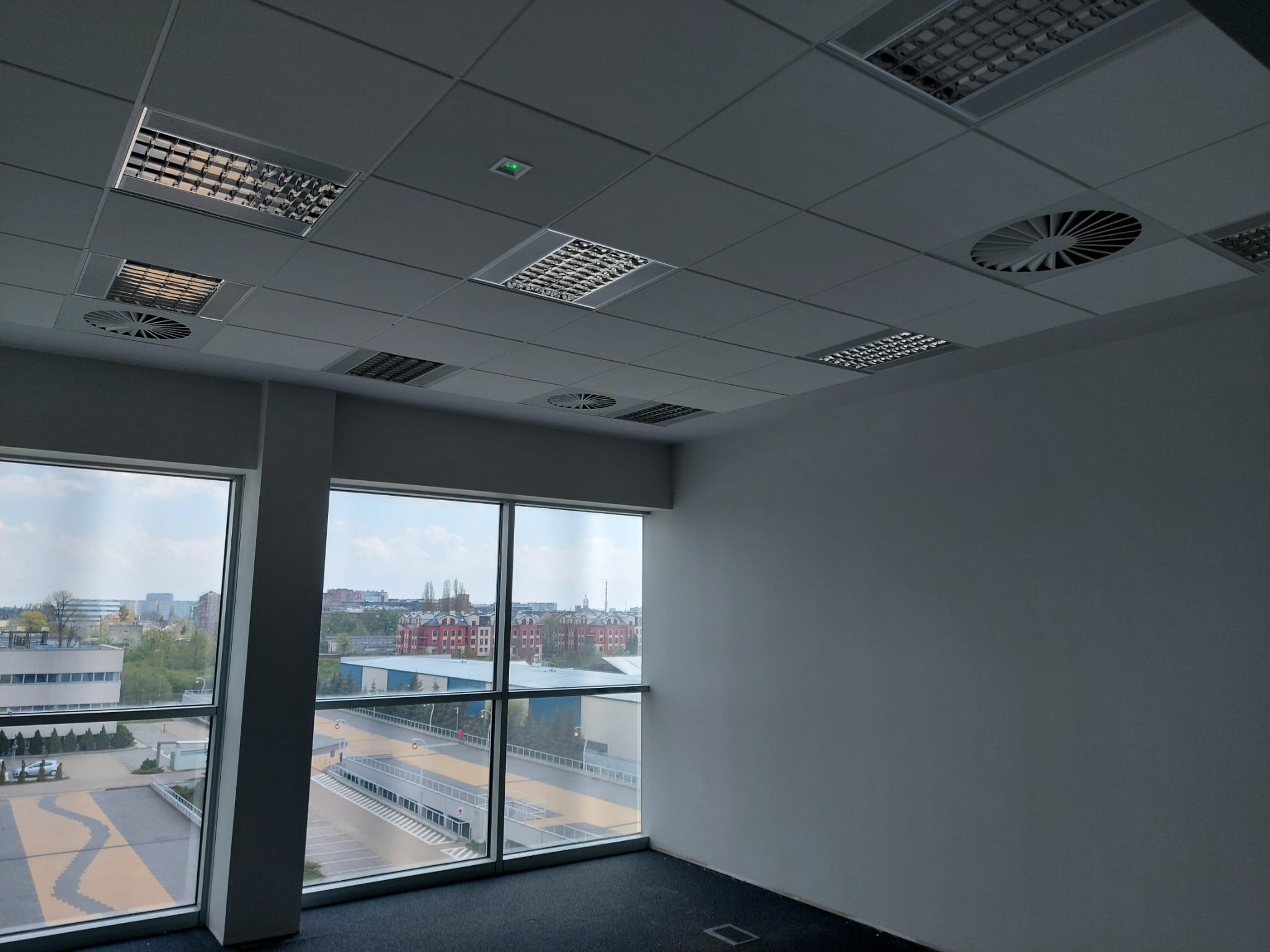 Wnętrze biura po malowaniu, widok z okna na miasto, sufit z rastrowymi lampami i kratkami wentylacyjnymi.