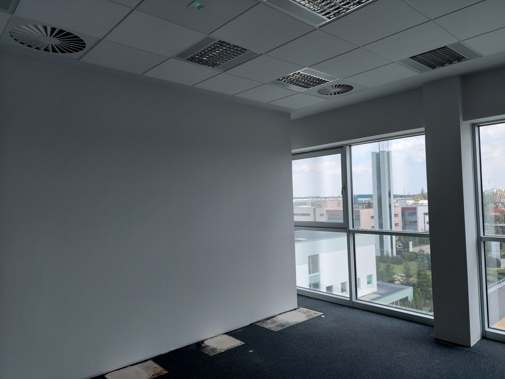 Biuro przed remontem: szara ściana, czarna wykładzina z widocznymi śladami po usuniętej podłodze, okna z widokiem na budynki biurowe.
