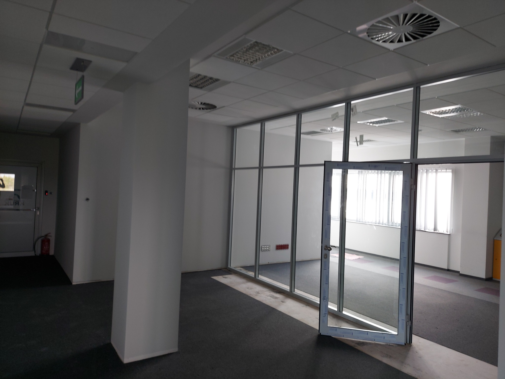 Wnętrze biura z przeszkloną ścianą działową i otwartymi drzwiami, widoczne instalacje pod sufitem, szara wykładzina podłogowa.