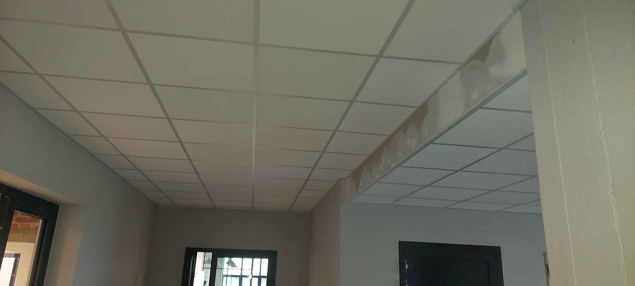 Kasetonowy sufit podwieszany z metalową konstrukcją, widoczne ślady szpachlowania na fragmencie konstrukcji, w tle widoczne drzwi i fragment pomieszczenia.