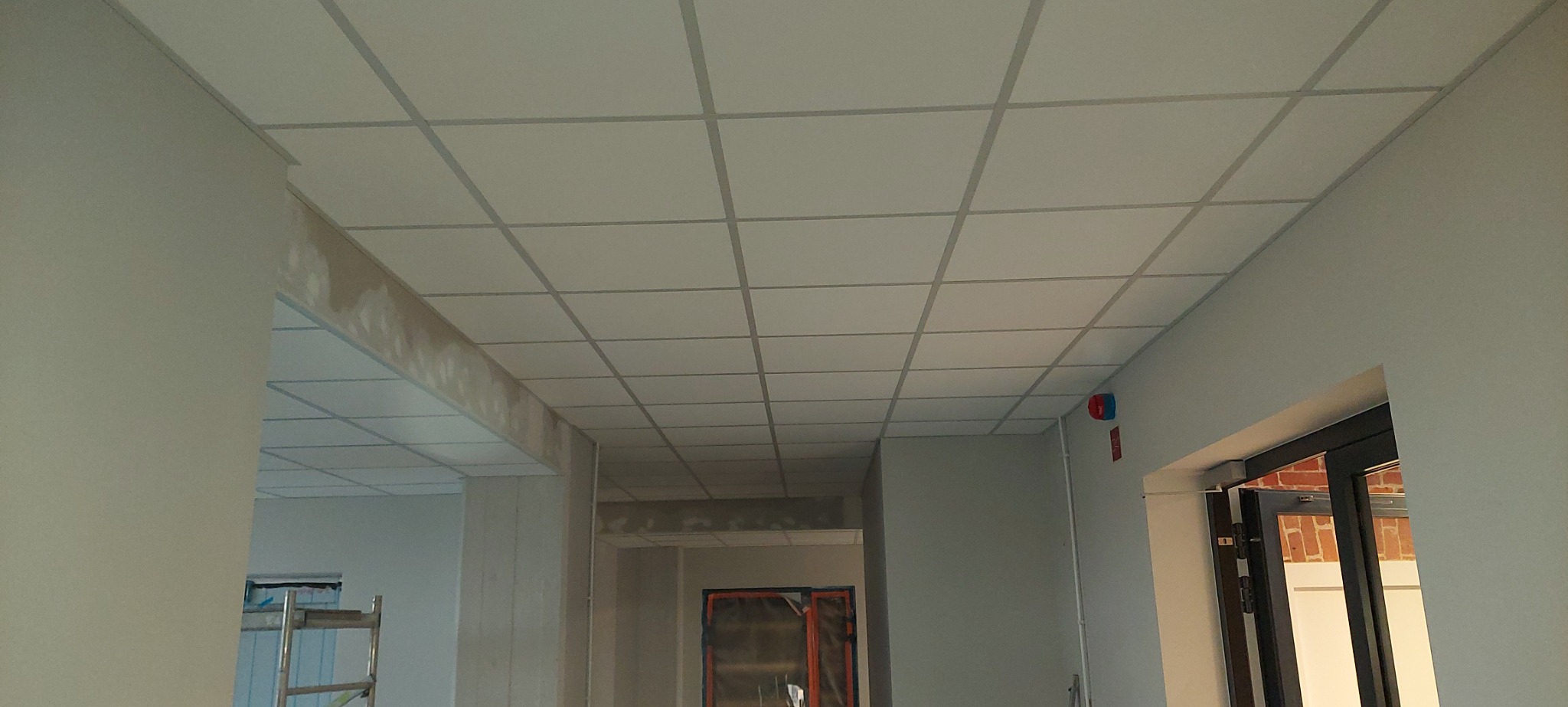Sufit kasetonowy w trakcie montażu w biurze, widoczne niedoskonałości na ścianach i sufitach, w tle drzwi wejściowe.