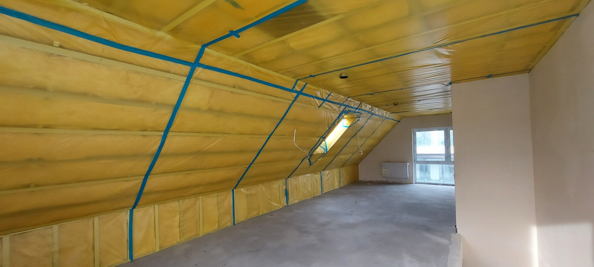 Poddasze zabezpieczone folią i taśmą malarską przed dalszymi pracami wykończeniowymi, widoczny fragment okna dachowego i grzejnik na ścianie.