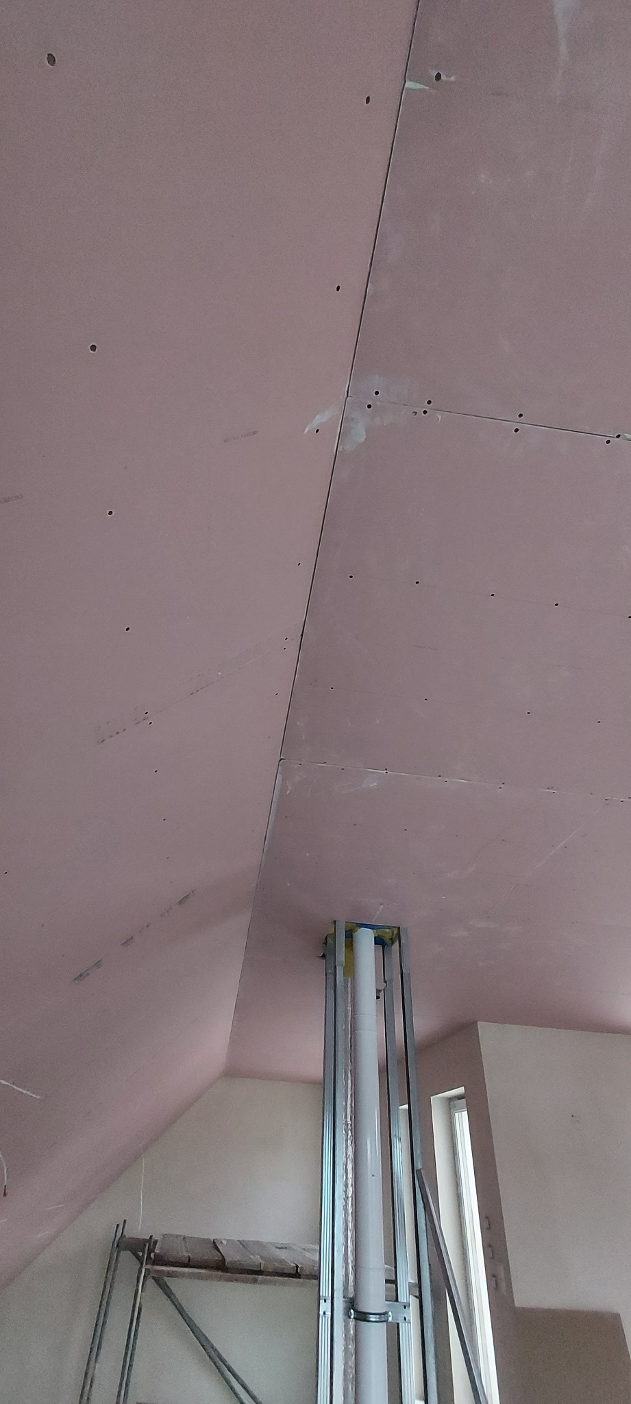 Montaż płyt kartonowo-gipsowych na poddaszu, widoczne łączenia płyt, metalowa konstrukcja wokół rury wentylacyjnej, fragment rusztowania.