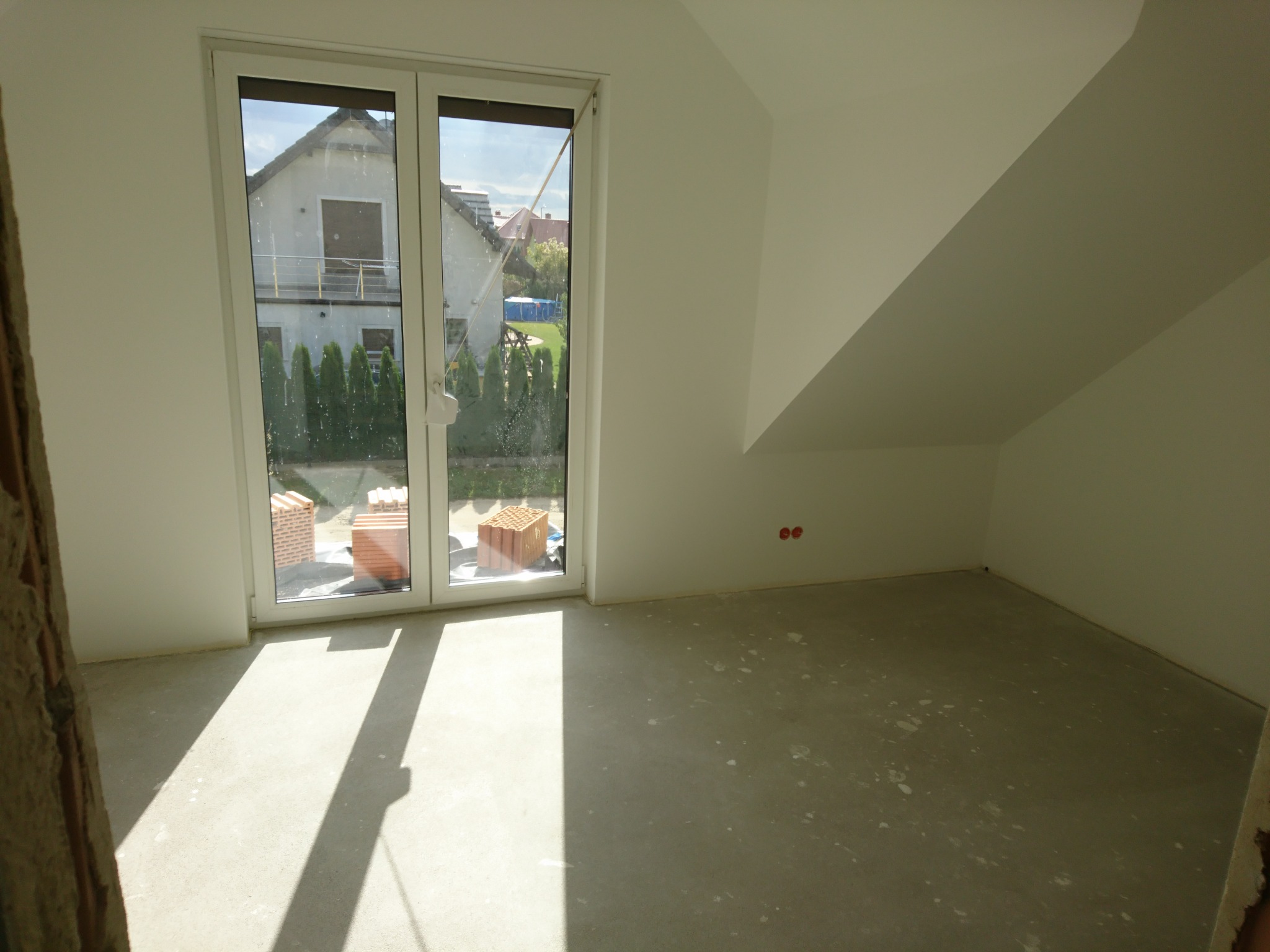 Pokój na poddaszu w trakcie prac wykończeniowych: surowy betonowy podkład podłogowy, białe ściany z widocznymi punktami elektrycznymi, okno balkonowe z widokiem na dom i ogród, cegły budowlane za...