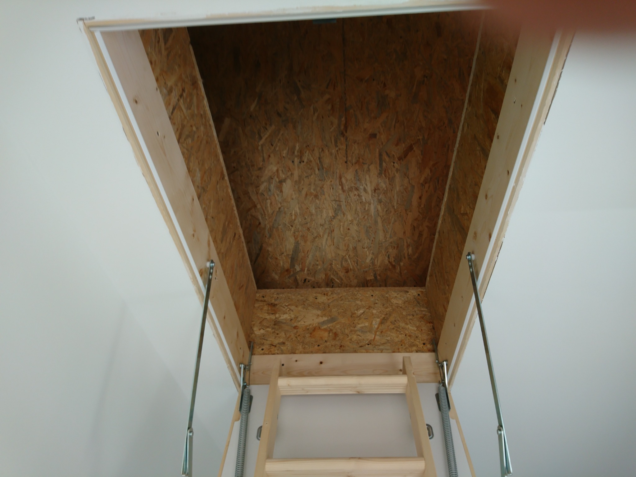 Widok od dołu na drewnianą konstrukcję otworu strychowego z zamontowaną składaną drabinką, widoczne sprężyny i metalowe elementy mocujące.